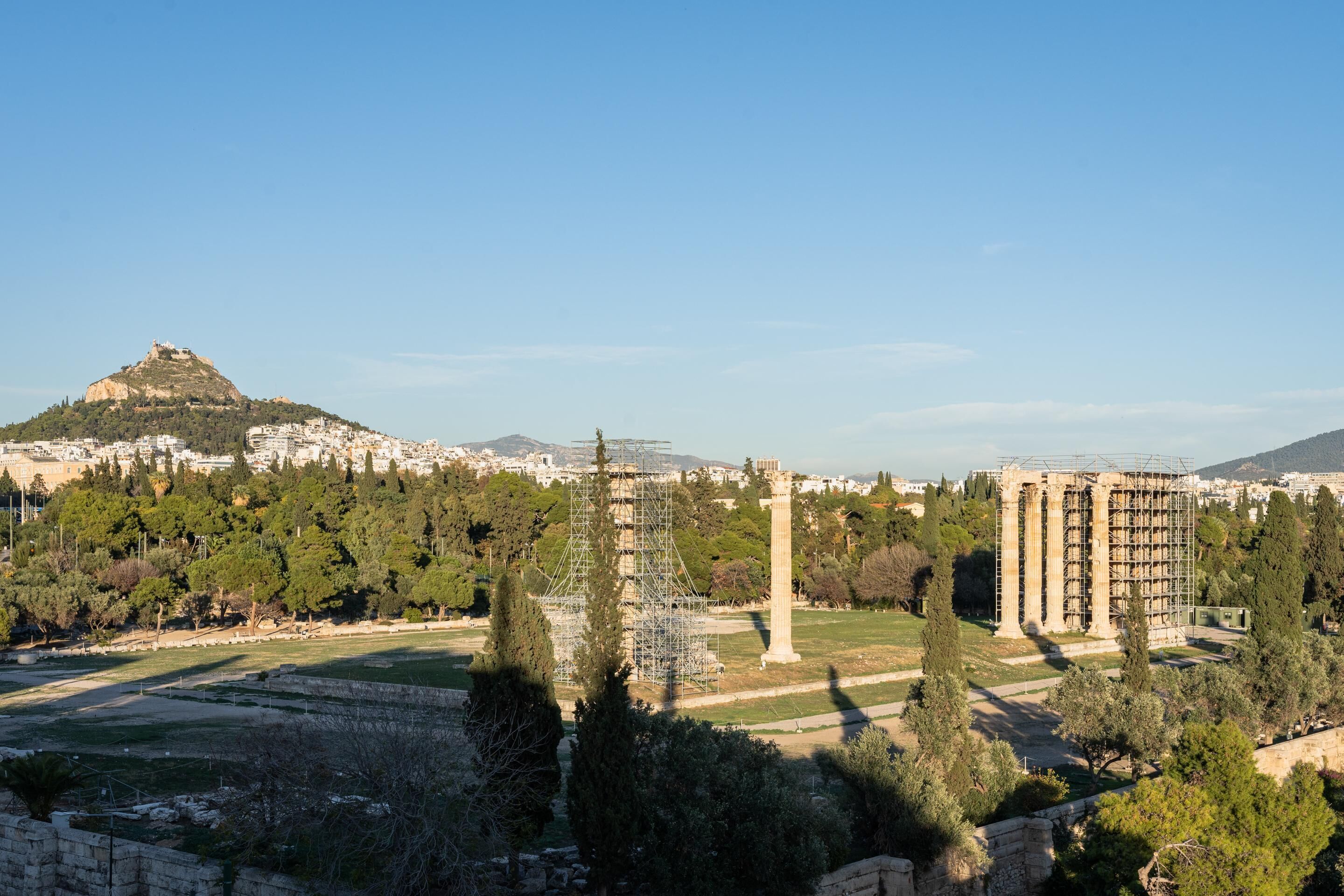 A historical site with ancient ruins, including columns and a temple, surrounded by trees and a hill in the background.