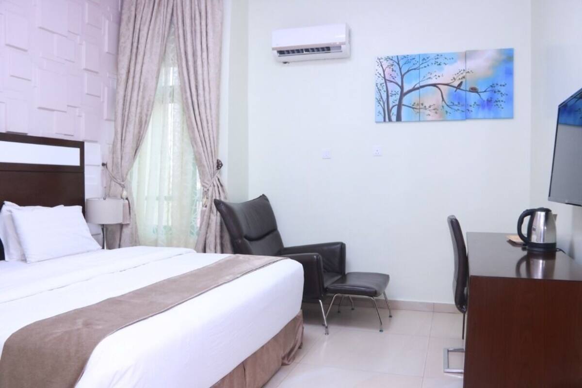 A hotel room with a bed, a desk, a chair, an air conditioner, and a painting on the wall.