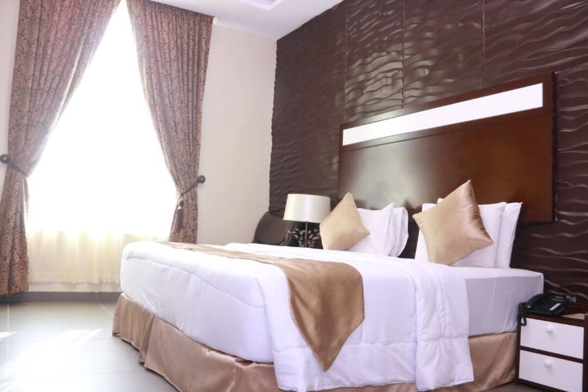 A hotel room with a large bed, a wooden headboard, a bedside table with a lamp, and a window with curtains.