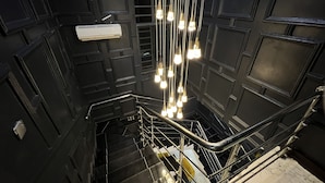Una escalera con barandilla de metal, paredes negras con patrones geométricos y lámparas colgantes.