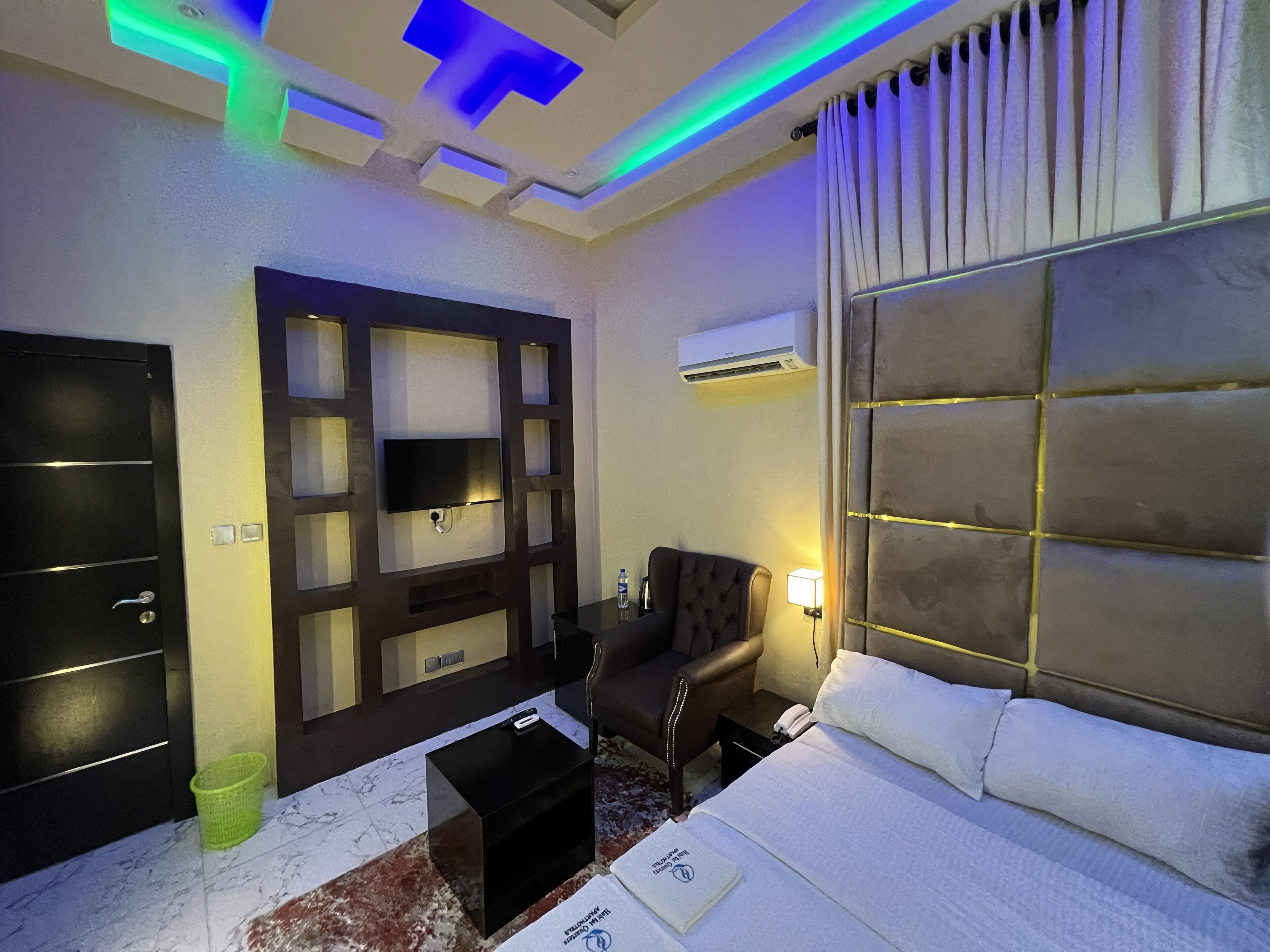 A hotel room with a bed, a TV mounted on a wall, a chair, a small table, and a trash bin.