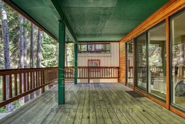 A wooden deck with a green railing, large windows, and a view of trees.