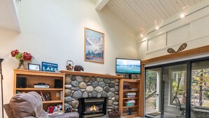 A cozy living room with a stone fireplace, a television, and a view of a forested area through a sliding glass door. - Cozy three-bedroom condo w summer pool (FP1-15) (Incline Village)