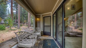 A covered porch with two chairs and a bench, a door leading inside, and a view of a forested area. - Cozy three-bedroom condo w summer pool (FP1-15) (Incline Village)
