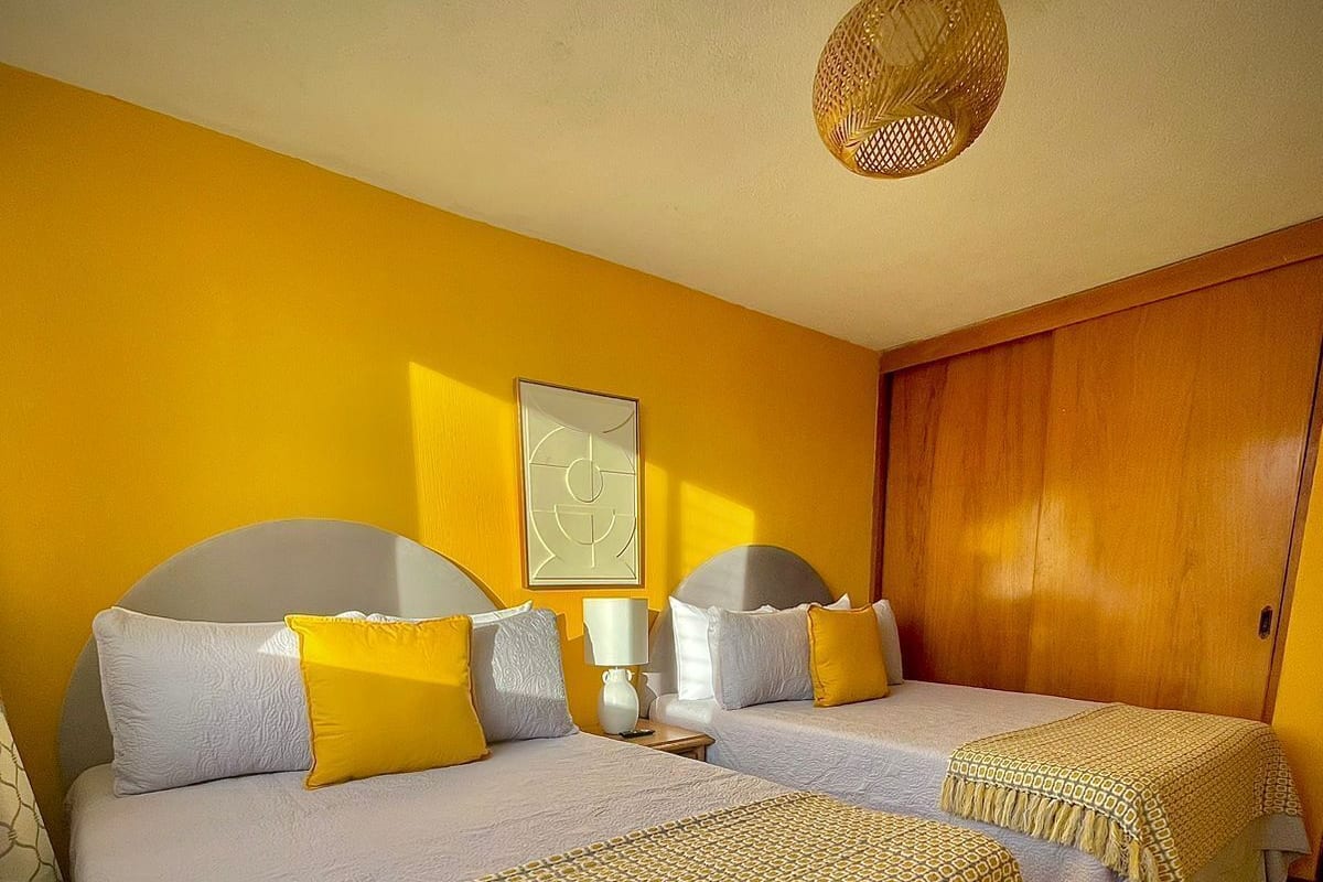 Ein Hotelzimmer mit zwei Betten, einem Holzschrank, einer gewobenen Deckenleuchte und einem gerahmten abstrakten Kunstwerk an der Wand.