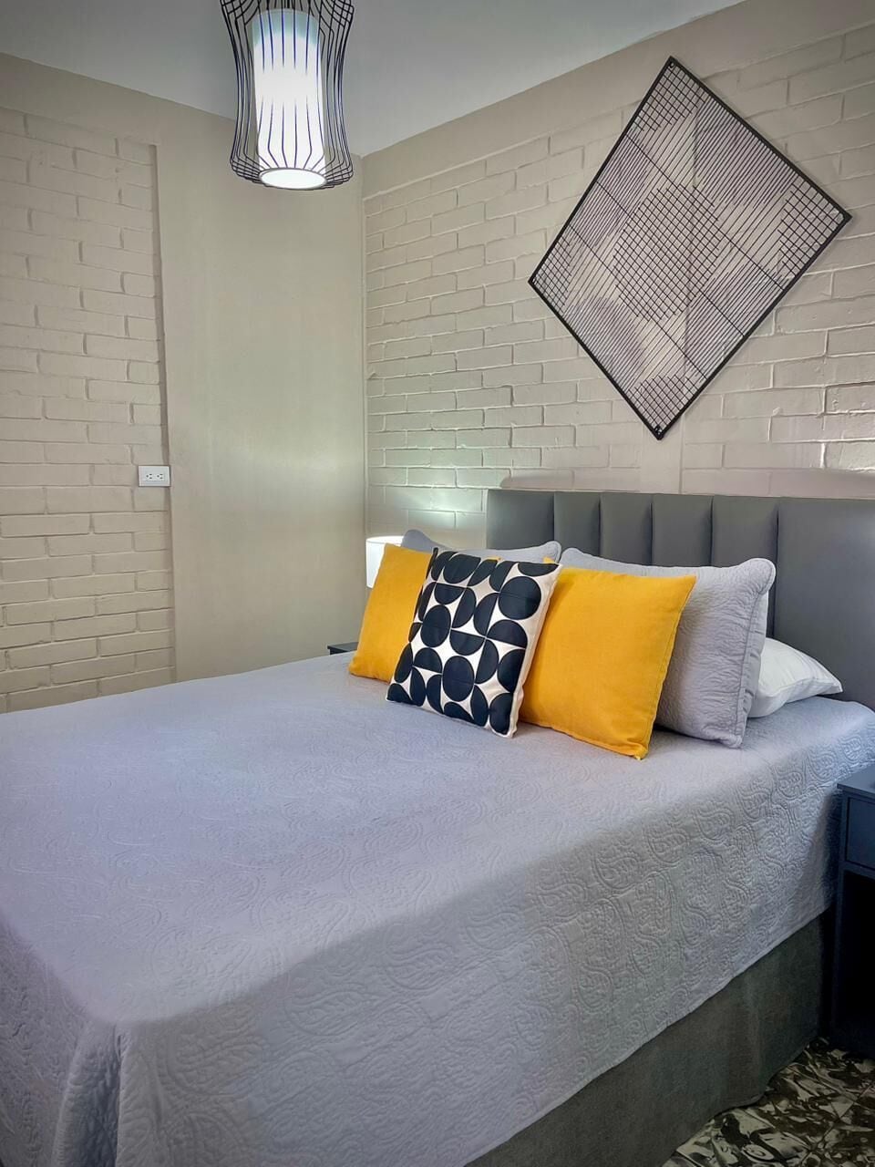 Una cama con cabecera gris, colcha blanca con textura y dos almohadas de colores sólidos y diseño geométrico.