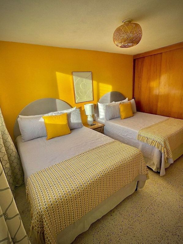 A hotel room with two beds, a wooden wardrobe, a bedside table with a lamp, and a wall-mounted artwork.