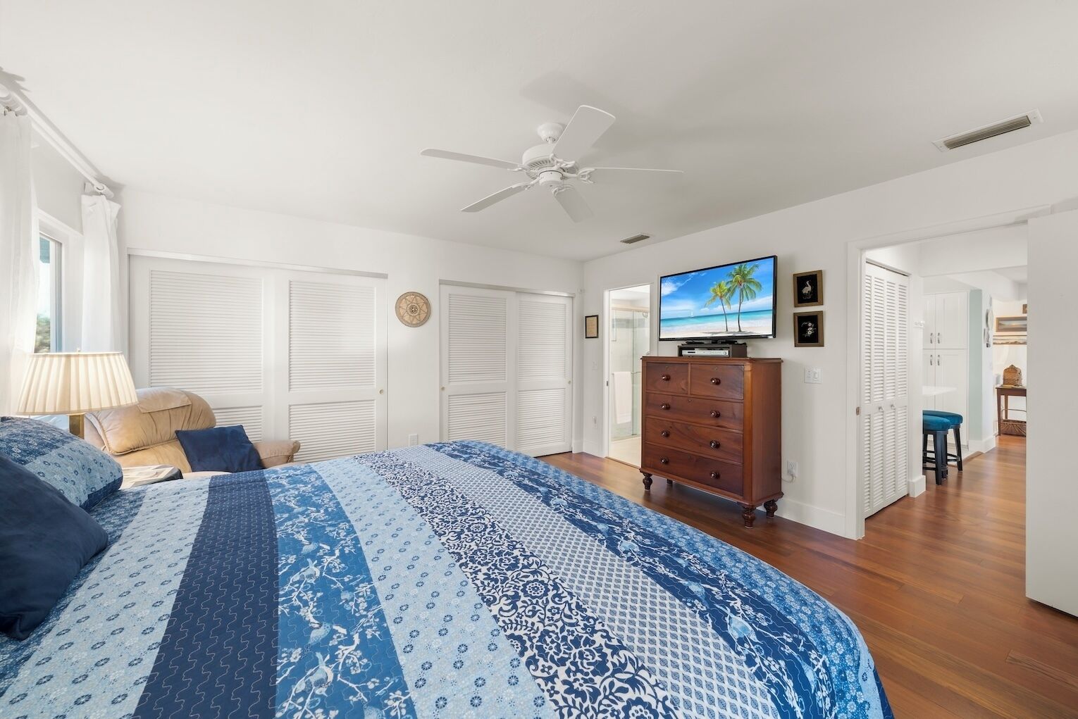 A bedroom with a large bed, a dresser, a ceiling fan, a TV, and a view of the beach through the window.