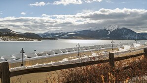 A lakeside view with a wooden fence, snow-covered ground, and mountains in the background. - Walk to the Dillon Amphitheater, Marina and more! (Dillon)