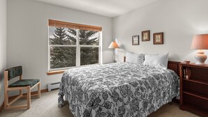 Ein Schlafzimmer mit einem großen Bett, einem Fenster mit Jalousien, einem Sessel, einem Nachttisch mit Lampe und gerahmten Bildern an der Wand.