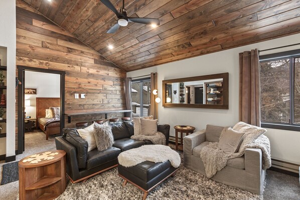 A cozy living room with a wooden ceiling, a black leather sofa, a grey armchair, and a small wooden side table.