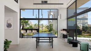 Une table de ping-pong dans une pièce moderne, dotée de grandes fenêtres, de chaises et d’une petite table.