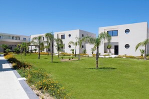 A modern building with a flat roof, circular windows, and a well-maintained lawn with palm trees and yellow flowers. - Trapezia Luxury Living - Opal (Afantou)