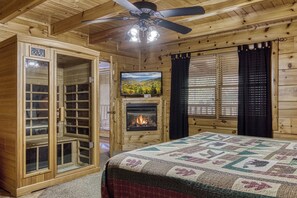 Interior de una cabaña de troncos con cama, chimenea, televisor y ventilador de techo.