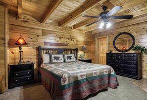 Un dormitorio acogedor con techo de madera, una cama con colcha, una mesita de noche con lámpara, un aparador y un ventilador de techo.