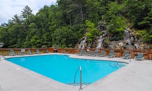 A rectangular swimming pool with a wooden deck and lounge chairs, surrounded by a wooden fence and a rocky waterfall backdrop.