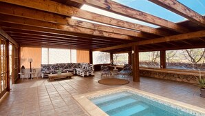 Una piscina interior con suelo de baldosas, vigas de madera y una zona de estar con sofá y sillones.