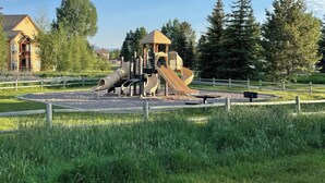 Un parque infantil con toboganes y estructuras para escalar, rodeado por una valla y césped, con casas y árboles al fondo.