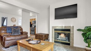 Una sala de estar con un sofá de cuero, una mesa de centro, una chimenea, un televisor montado en la pared y una cocina al fondo.