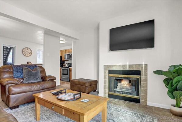 A living room with a leather sofa, a coffee table, a fireplace, a TV mounted on the wall, and a kitchen in the background.