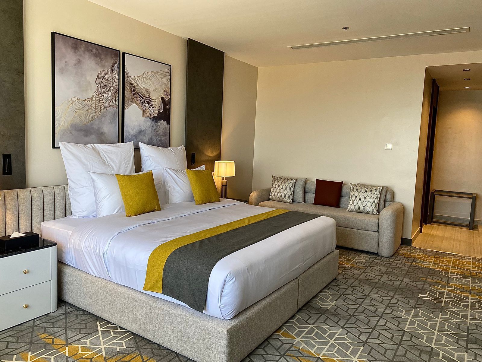 Una habitación de hotel moderna con una cama grande, un sofá y una mesita de noche. Hay cuadros abstractos en la pared y una lámpara sobre el sofá.