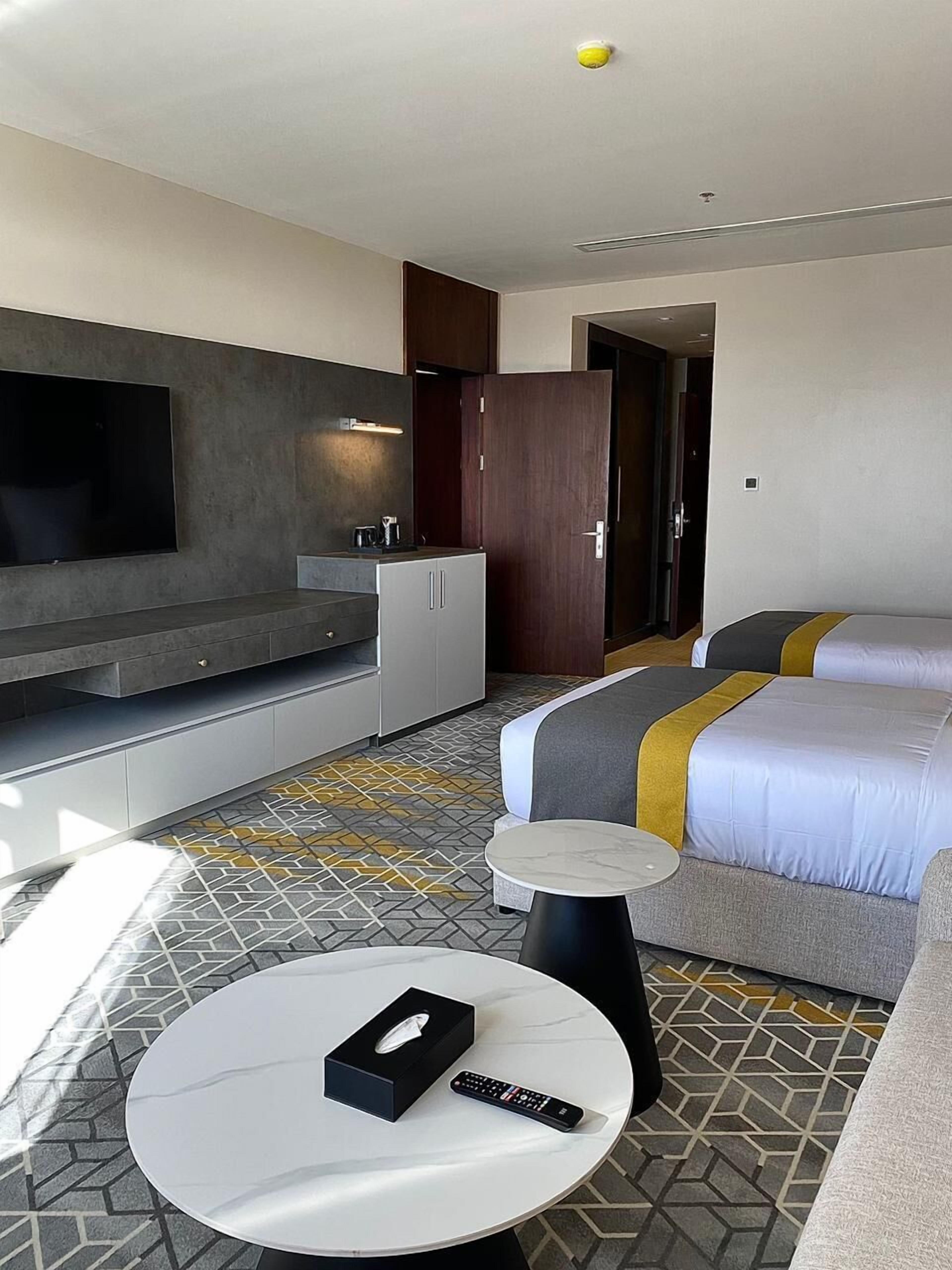 Una camera d'albergo moderna con televisione a schermo piatto, un letto con copriletto grigio e giallo, un tavolino rotondo con un telecomando e un pavimento decorato.