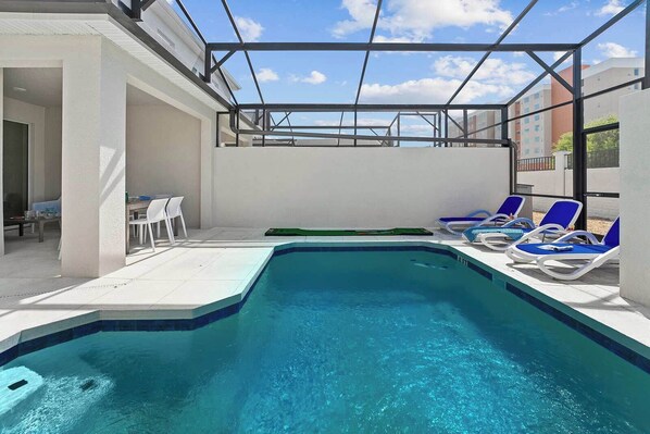 A swimming pool with blue water, surrounded by white walls and a glass ceiling. There are lounge chairs and a table with chairs on the poolside.