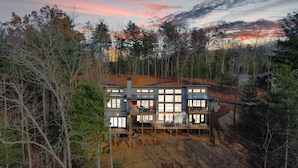 A modern two-story house with large windows, a deck, and a chimney, surrounded by trees.