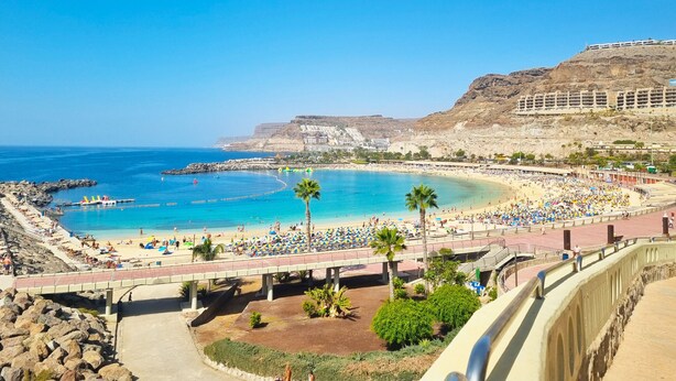 A beach with a large crowd, a clear blue sky, and rocky cliffs in the background.
