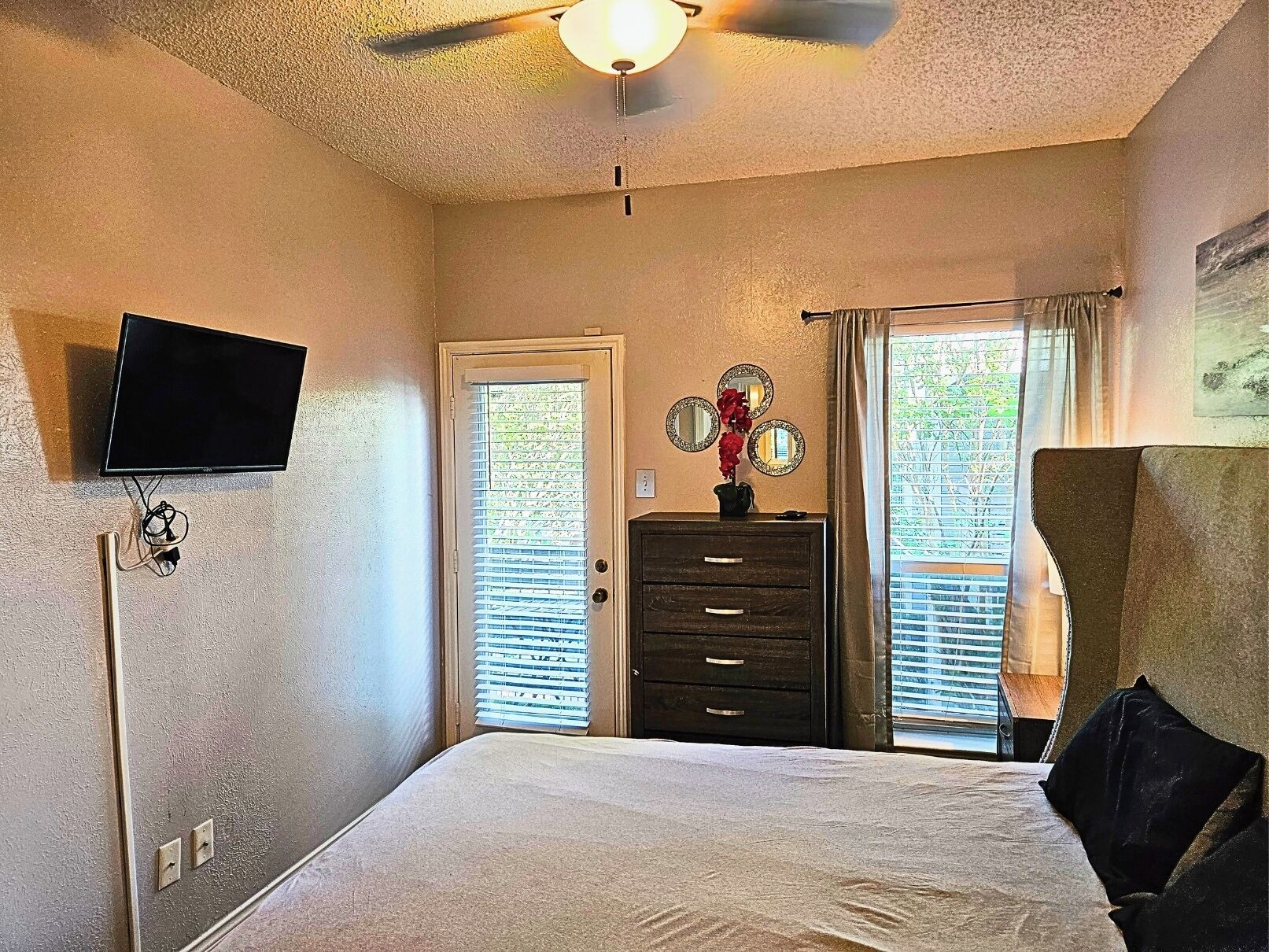 A bedroom with a bed, a dresser, a television mounted on the wall, and a ceiling fan.
