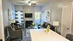 A modern living room with a fireplace, a ceiling fan, and a flat-screen TV mounted on the wall. - I30 - Apt|Patio|11mi to Convention Ctr|Sleeps 2 (Irving)