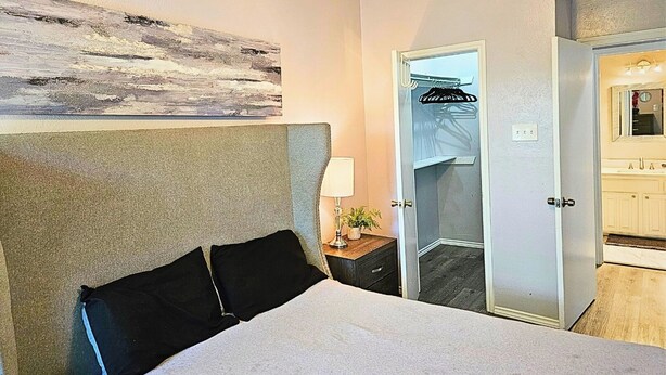 A hotel room with a bed, a nightstand, a lamp, a mirror, a door, and a wall with a painting. - I30 - Apt|Patio|11mi to Convention Ctr|Sleeps 2 (Irving)