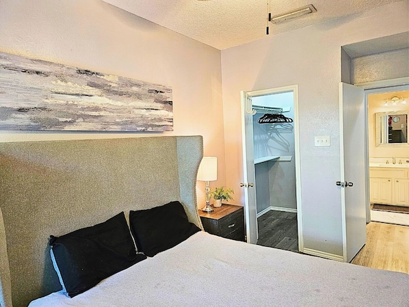 A hotel room with a bed, a nightstand, a lamp, a mirror, a door, and a wall with a painting.