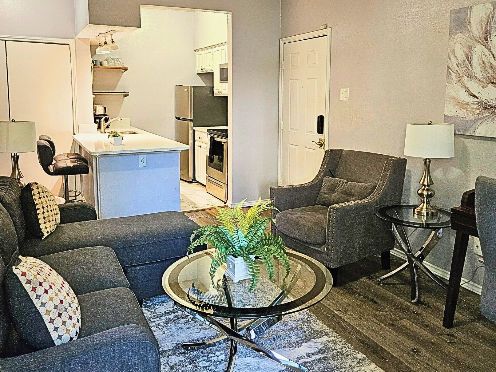 A living room with a sofa, armchair, and a coffee table with a plant. There is a kitchen area in the background with cabinets and appliances.