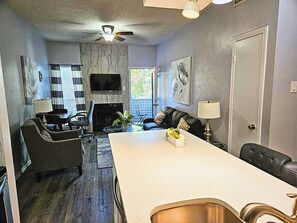 A modern living room with a fireplace, a flat-screen TV, and a dining area with a kitchen island. - I30 - Apt|Patio|11mi to Convention Ctr|Sleeps 2 (Irving)
