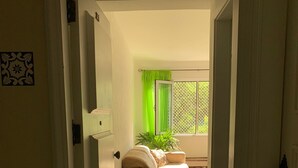 Un couloir mène à une chambre à coucher qui contient un lit, une fenêtre avec des rideaux verts et une plante en pot.