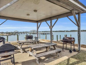 An outdoor patio with picnic tables, a grill, and a view of a dock and water. - Sea Oats J104 Lagoon Retreat w / Pier & Boat Ramp (Gulf Shores)