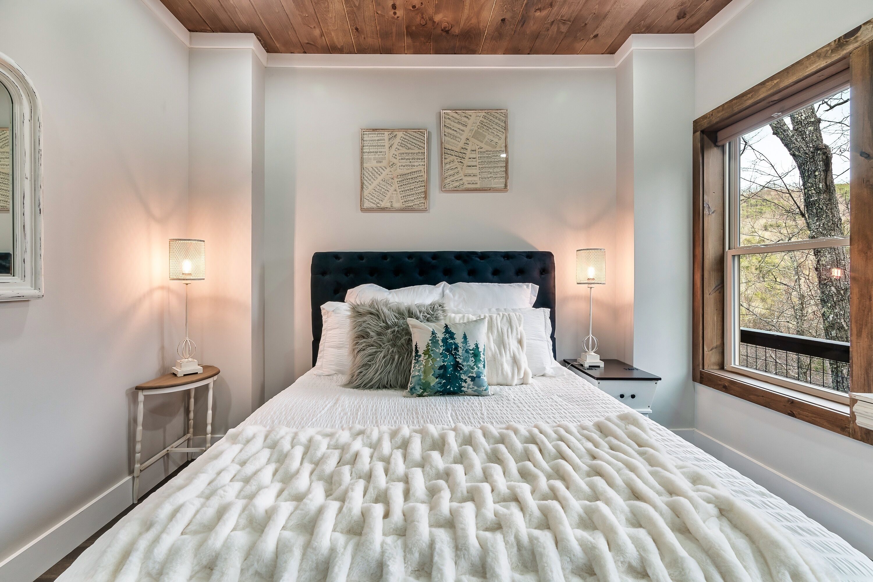 Een slaapkamer met een groot bed, twee wandlampen, een raam met uitzicht op bomen en een houten plafond.