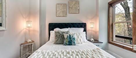 Een slaapkamer met een groot bed, twee wandlampen, een raam met uitzicht op bomen en een houten plafond.