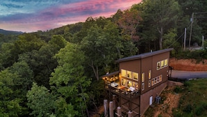 A modern, two-story wooden cabin with a balcony, surrounded by dense forest. - Fall Getaway! Hot Tub! Mountain Views! (Gatlinburg)