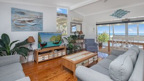 A modern living room with a wooden floor, grey sofas, a wooden coffee table, a TV, and a large window with a view of the sea. - Paradise on Partridge (Port Macquarie)