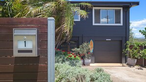 A modern two-story house with a dark exterior, a garage door, and a well-maintained garden with various plants and flowers. - Paradise on Partridge (Port Macquarie)