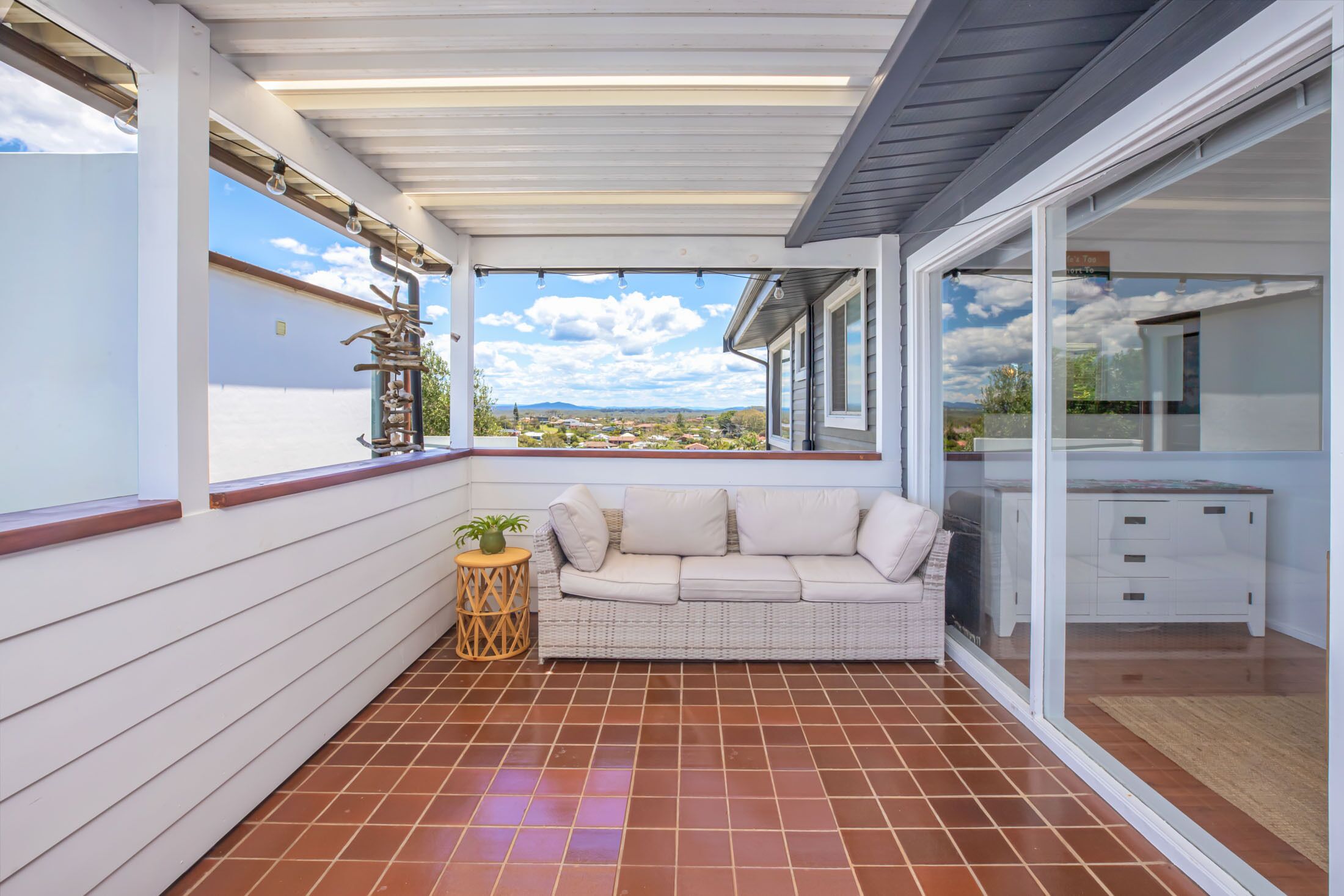 Een overdekt balkon met een witte bank, een klein rond tafeltje met een plant en uitzicht op de omgeving.