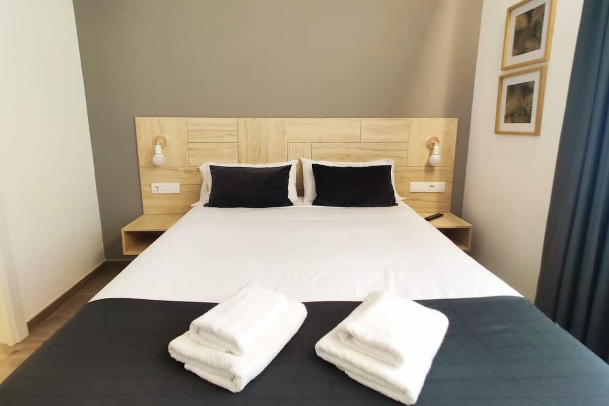 Una cama bien tendida con sábanas blancas y dos almohadas negras, un cabecero de madera, mesitas de noche con lámparas y una cortina azul.