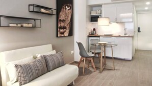 A modern living room with a white sofa, a coffee table, and a dining area with two chairs. There is a kitchen area with white cabinets and a microwave. A large framed artwork is on the wall, and there is a ceiling light fixture. - GLOBAL Apartments & Rooms (L'Hospitalet de Llobregat)