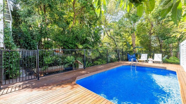 Une piscine dotée d’une terrasse en bois, entourée d’une végétation luxuriante et d’une clôture en métal.