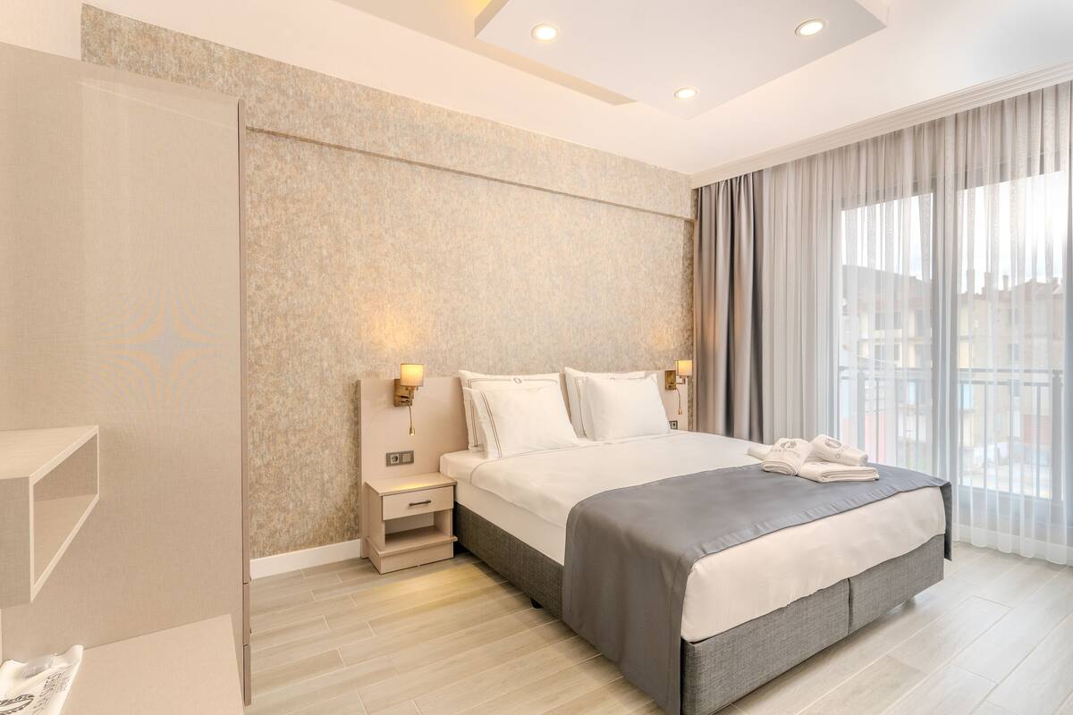 A modern hotel room with a large bed, bedside tables, and a view of the city through large windows.