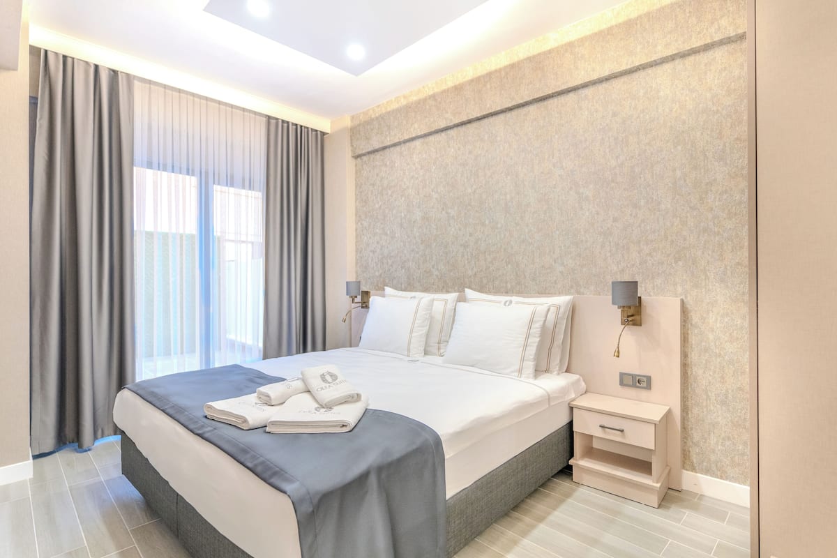 A modern hotel room with a large bed, a bedside table with a lamp, and a view of the outdoors through the window.