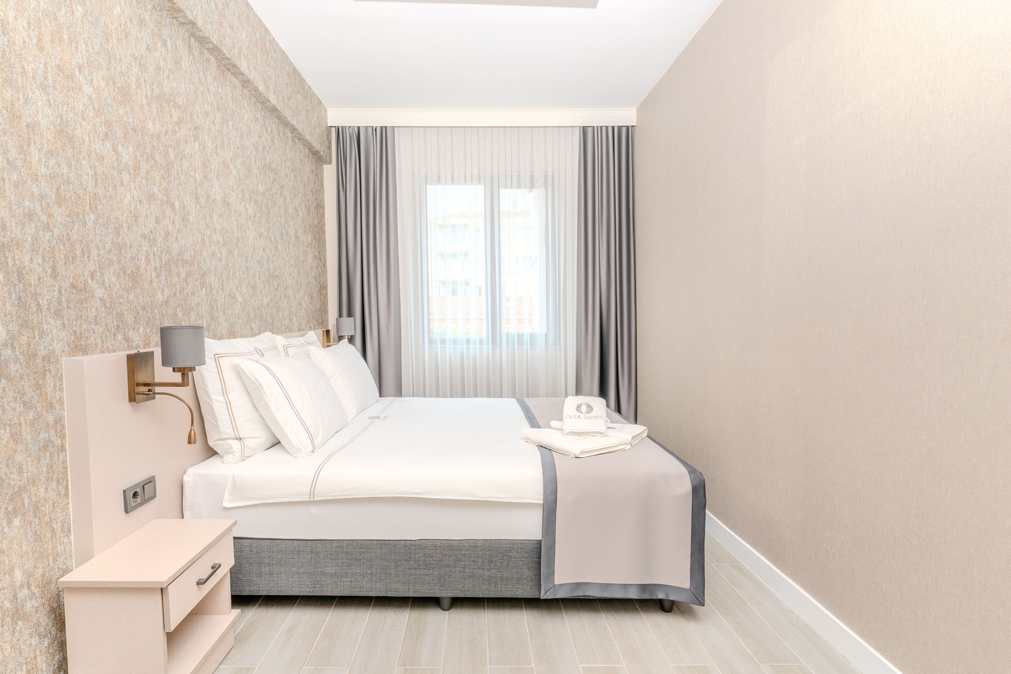 Ein modernes Hotelzimmer mit einem ordentlich bezogenen Bett, einem Nachttisch mit Lampe und einem großen Fenster mit Vorhängen.