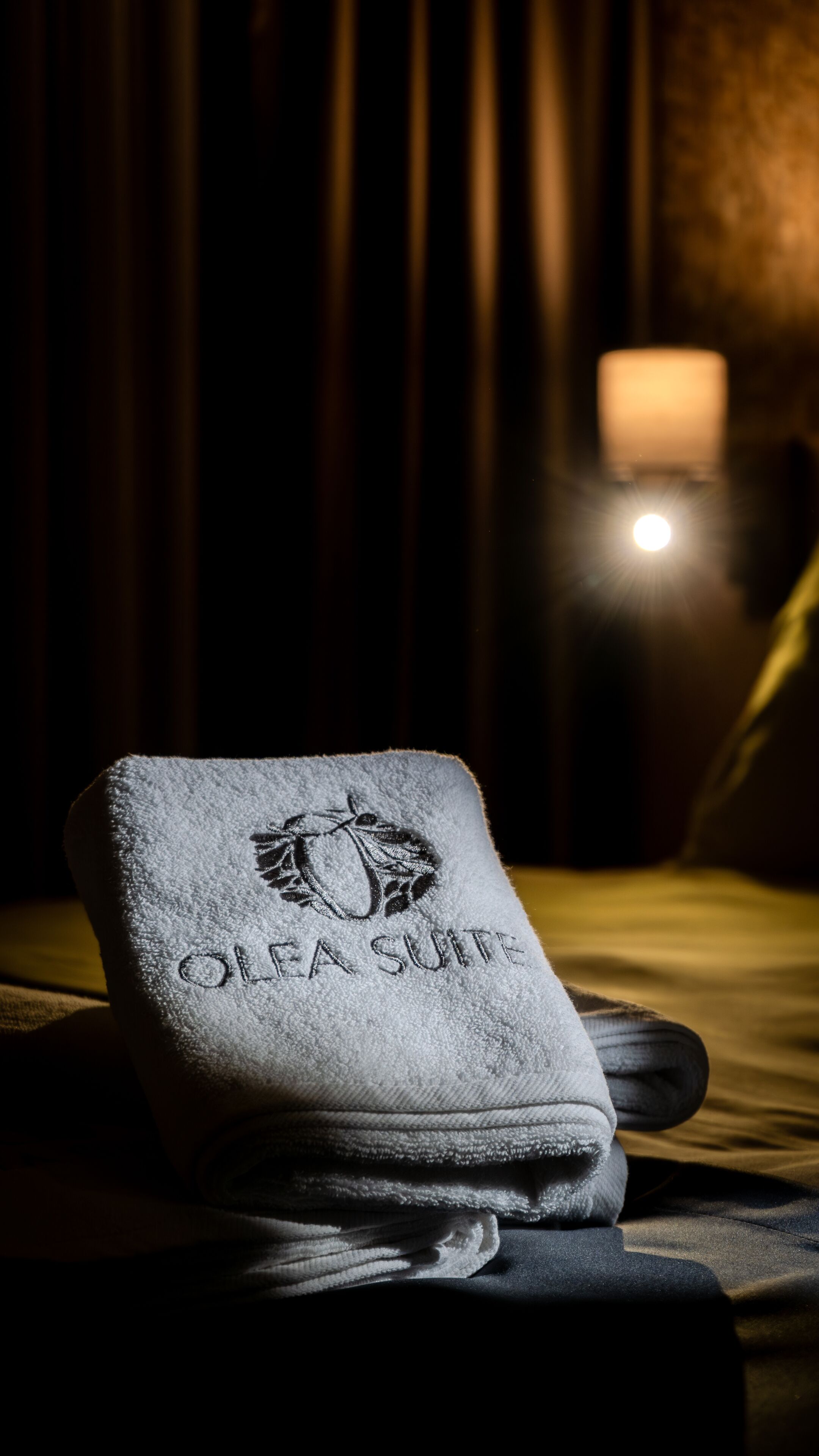Una toalla doblada con el bordado 'Olea Suite', sobre una cama en una habitación con poca luz, con una mesita de noche y una lámpara al fondo.